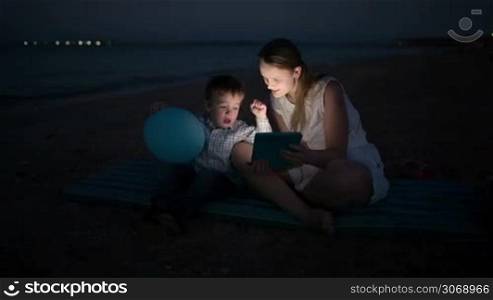 Mother and her little son with touchpad on the beach at night