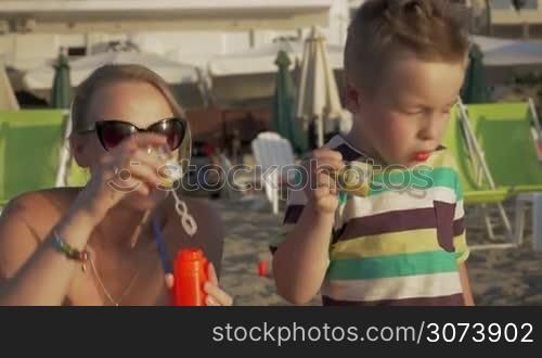 Mother and her son are resting on the beach in windy day and blowing soap bubbles.