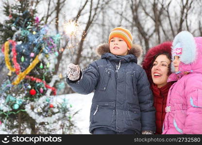 mother and her two children is standing near christmass tree and looking at bengal light in boy&acute;s hand.