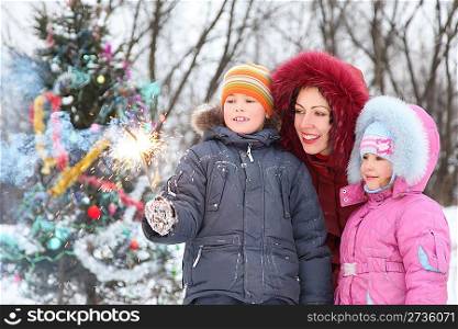 mother and her two children is standing near christmass tree and looking at bengal light in boy&acute;s hand. christmass tree in out of focus.