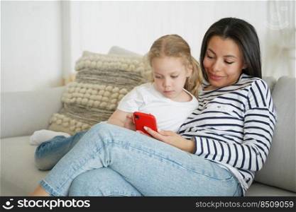 Mother and little kid girl playing online game on mobile phone, sitting on couch. Mom and daughter using learning app on smartphone, watching cartoons, resting on sofa at home together.. Mother and little daughter watching cartoons on smartphone, resting on sofa at home together