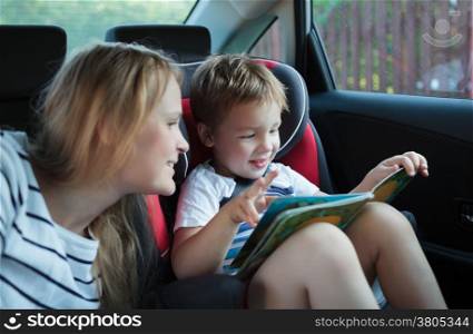 Mother and little son in the car. Woman watching boy looking through the book. Kid sitting in child safety seat