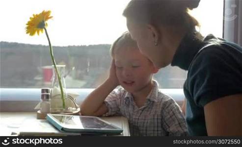 Mother and little son sitting at the table by the window in train. Child using tablet computer and they talking