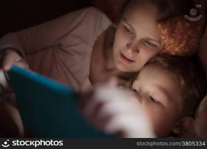 Mother and little son watching video or playing on touchpad lying in bed at night