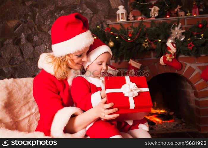 Mother and son are sitting near fireplace and christmas tree. Family look into a gift box.
