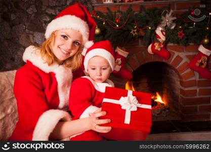 Mother and son are sitting near fireplace and christmas tree. Family look into a gift box.. Mother and son near Christmas fireplace