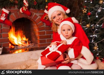 Mother and son are sitting near fireplace and christmas tree. Family look into a gift box.. Mother and son near Christmas fireplace