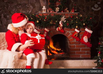 Mother and son are sitting near fireplace and christmas tree. Family look into a gift box.. Mother and son near Christmas fireplace