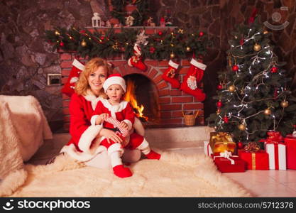 Mother and son are sitting near fireplace and christmas tree with gift boxes.