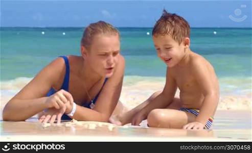 Mother and son at the seaside. Little smiling boy watching mom drawing on sand, water washing the drawing away