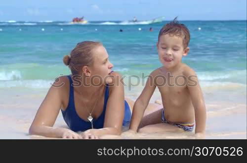 Mother and son at the seaside playing, little smiling boy watching mom drawing on sand