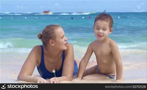 Mother and son at the seaside playing, little smiling boy watching mom drawing on sand