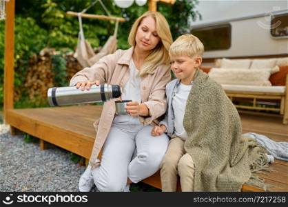 Mother and son drink tea from a thermos at the trailer, summer camping. Family with children travel in camp car, nature and forest on background. Campsite adventure, travelling lifestyle. Mother and son drink tea from a thermos at trailer
