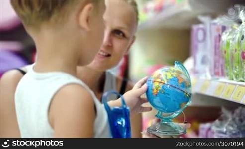 Mother and son in the shop. Woman holding small globe and showing countries, boy rotating it quickly