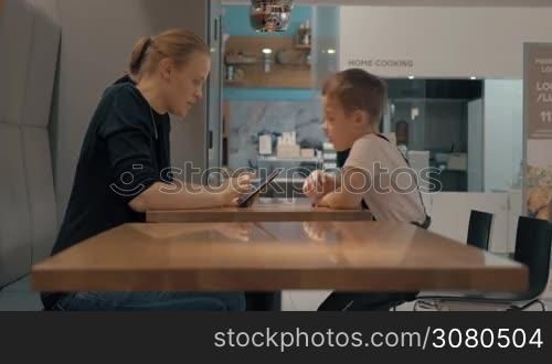 Mother and son sitting at the table in cafe and using mobile. Child asking mom to find something for him in the internet