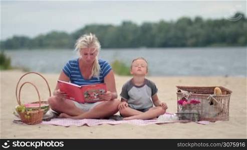 Mother and son sitting on colorful blanket and reading book during picnic on the beach. Family spending time together outdoor.