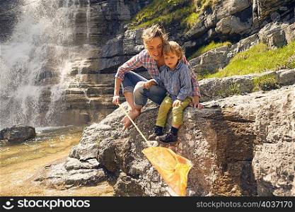 Mother and son, sitting on rocks by waterfall, fishing with net
