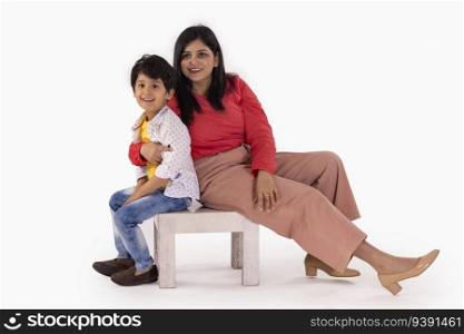 Mother and son sitting together on a wooden stool against white background