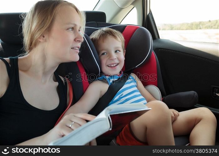 Mother and son traveling by car. Woman reading a book to the kid sitting in child safety seat