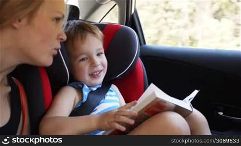 Mother and son traveling by car. Woman talking to boy sitting in child safety seat and holding a book