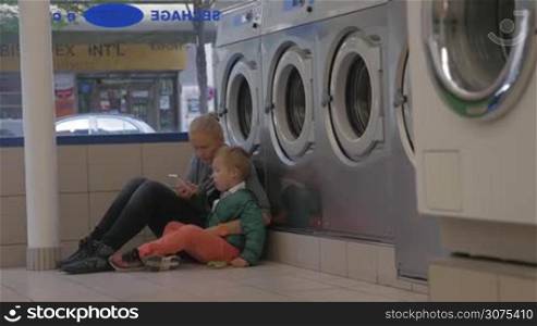 Mother and son waiting in the laundry. They sitting on the floor near the washing machines and using smart phone