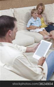 Mother and son watch television while man reads a digital book