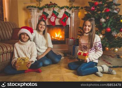 Mother and two daughter sitting with Christmas gifts on floor next to burning fireplace