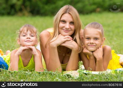 Mother and two daughters lie on the green grass and propping up hands a head looked in the picture