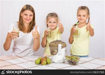 Mother and two daughters show a thumbs up freshly prepared juice. Young beautiful mother and two daughters sitting at a table squeezed juice from pears and grapes with a juicer