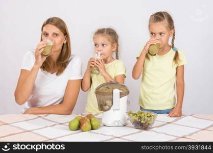 Mother and two daughters taste the freshly prepared juice from pears and grapes. Young beautiful mother and two daughters sitting at a table squeezed juice from pears and grapes with a juicer