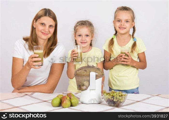 Mother and two daughters with glasses of freshly made juice from the pears and grapes. Young beautiful mother and two daughters sitting at a table squeezed juice from pears and grapes with a juicer