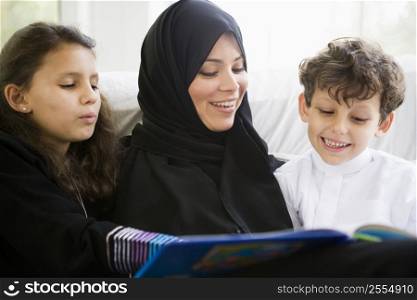 Mother and two young children in living room reading book and smiling (high key/selective focus)