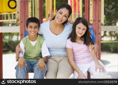Mother and two young children sitting on playground structure smiling (selective focus)