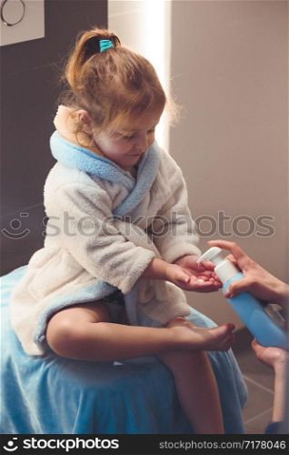 Mother applying moisturizing cream on her daughter&rsquo;s legs after bath. Mom caring about her child. Girl sitting in bathroom, wearing bathrobe