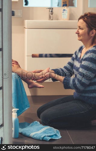 Mother applying moisturizing cream on her daughter&rsquo;s legs after bath. Mom caring about her child. Girl sitting in bathroom, wearing bathrobe