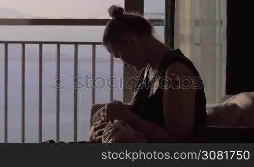 Mother breastfeeding baby daughter sitting in the arm-chair and looking at the sea through open balcony door. Mom and child bonding