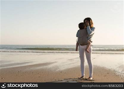 Mother carrying baby. Mother with ergobaby carrying toddler on the beach