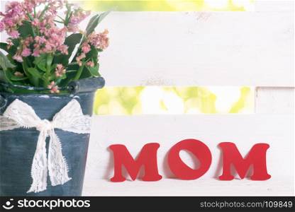 Mother day greeting card idea with the word mom written with red paper letters leaned against a white fence, near an old bucket full with flowers.