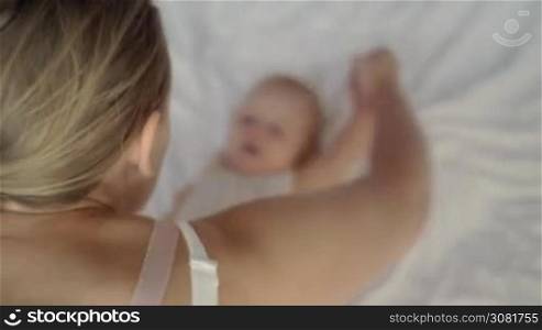 Mother doing gymnastics to baby daughter lying on bed. Mom and child doing hand exercises