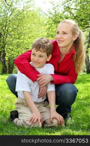 Mother embraces behind son sitting on laps on grass in park in spring