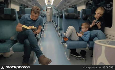 Mother, father and child having a train journey. Tired child using pad leaning on mother who browsing on smart phone. Dad prefers a book to electronics