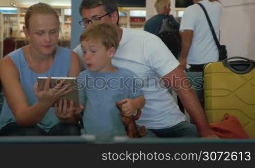 Mother, father and little son in the waiting-room of the airport. They using tablet computer to entertain the son