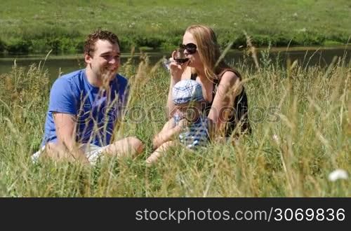 Mother, father and little son sitting on the grass near the small river. Woman drinking wine, boy eating. Family picnic on a sunny summer day