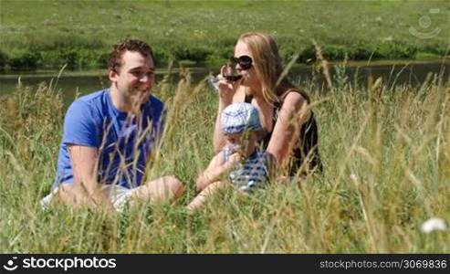 Mother, father and little son sitting on the grass near the small river. Woman drinking wine, boy eating. Family picnic on a sunny summer day