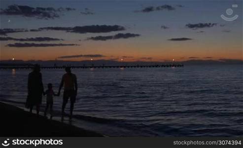 Mother, father and son holding hands and walking barefoot along the sea shore in the dusk. Black silhouettes with dark sea, sky and pier in background
