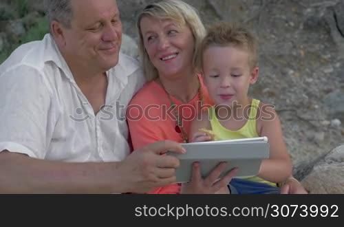 Mother,father and son watching video on pad while sitting on beach. Boy eating food, parents talking.