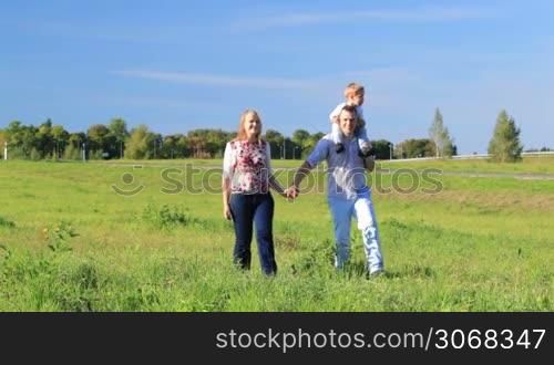 Mother, father and their little son walking in the countryside. Couple holding hands, man kissing his wife while boy sitting on father&acute;s shoulders.