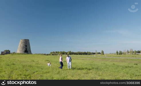 Mother, father and their little son walking on green grass in the countryside. Rural buildings and wood with clear blue sky in background.