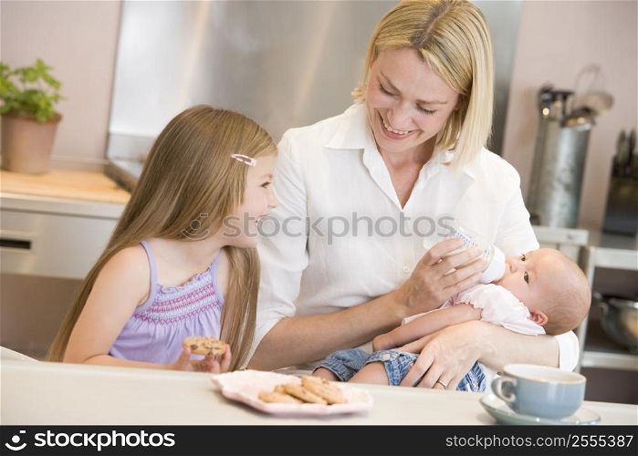 Mother feeding baby in kitchen with daughter eating cookies and smiling