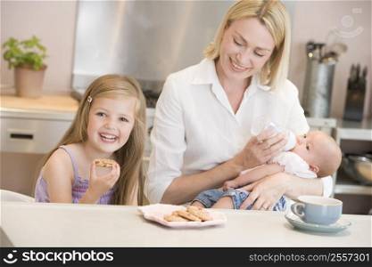 Mother feeding baby in kitchen with daughter eating cookies and smiling